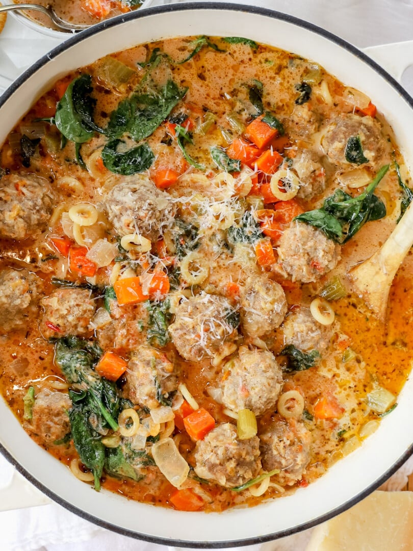 Overhead view of creamy meatball soup with pasta, meatballs, and spinach in a white pot.