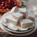 A side view of cranberry marshmallows garnished with fresh cranberries and rosemary on a stack of white plates.