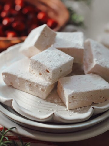 A side view of cranberry marshmallows garnished with fresh cranberries and rosemary on a stack of white plates.