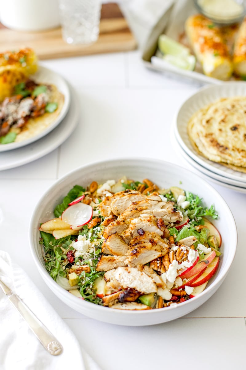 Harvest chicken salad with sliced chicken, apples, pecans, feta cheese, and avocado served with sourdough tortillas and strawberry avocado caprese salad in the background.
