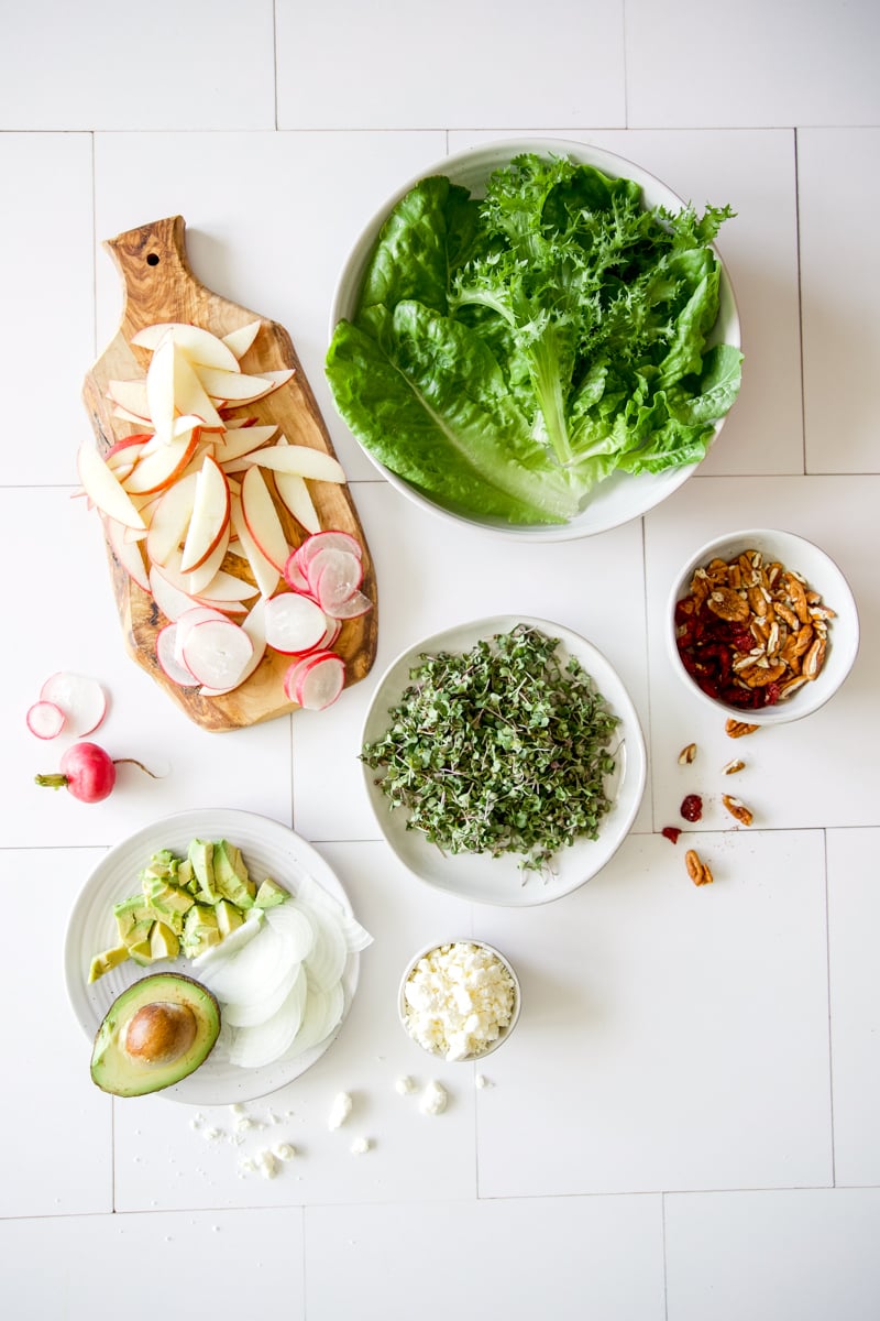 Ingredients for harvest chicken salad including lettuce, microgreens, apples, radishes, avocado, feta cheese, pecans, and dried cranberries arranged on a white table.