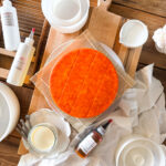 Beginner cheesemaking equipment including a homemade Colby cheese wheel, rennet, annatto, cheese molds, cultures, and curd cutting tools.