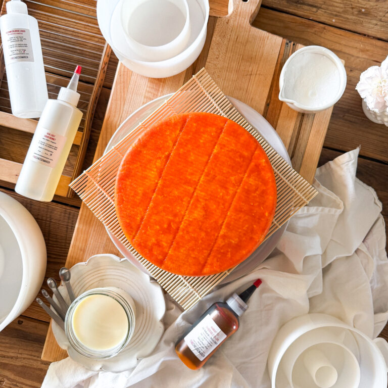 Beginner cheesemaking equipment including a homemade Colby cheese wheel, rennet, annatto, cheese molds, cultures, and curd cutting tools.