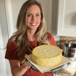 Meg holding homemade Gouda cheese wheel held in kitchen after pressing, made from fresh milk using traditional method.