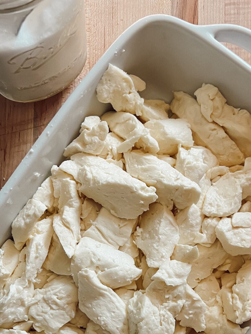 Fresh homemade cheddar cheese curds in a white baking dish on a wooden cutting board.