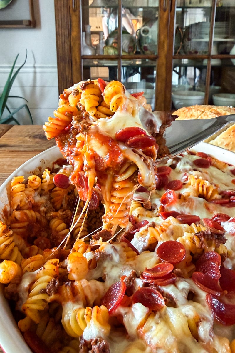 Cheesy pizza pasta bake made with ground beef or venison, rotini pasta, pepperoni, pizza sauce, and melted mozzarella cheese showing a spoon lifting a serving out with a long cheese pull.
